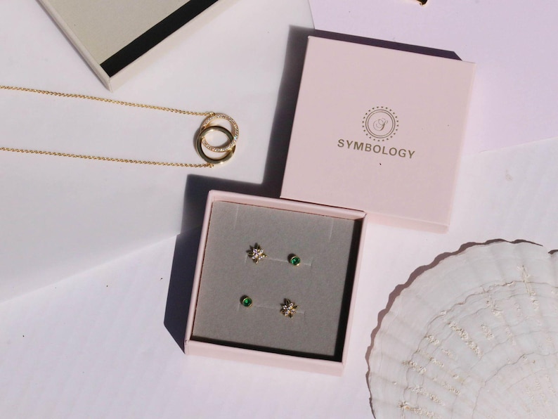 May include: A gold necklace with interlocking circles and a pair of stud earrings in a pink box. The earrings include a pair of star-shaped studs and a pair of green gemstone studs. The box is labeled "SYMBOLOLOGY".