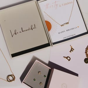 Può includere: Un assortimento di gioielli dorati, tra cui collane, bracciali e ciondoli, presentati in confezioni regalo. Una scatola recita "Life is beautiful", mentre un'altra mostra una collana con la parola "mamma". È visibile anche un braccialetto con un ciondolo a forma di lettera.