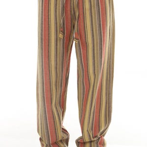 May include: Full-length trousers featuring a vertical striped pattern in shades of mustard yellow, burnt orange, and dark brown. The pants have an elasticized waistband with a matching fabric drawstring tie and a loose, relaxed fit throughout.