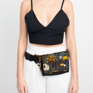 May include: A black crop top with spaghetti straps and a deep V-neck. A floral print fanny pack with a black buckle is worn around the waist. The fanny pack has a dark background with yellow and brown floral patterns.