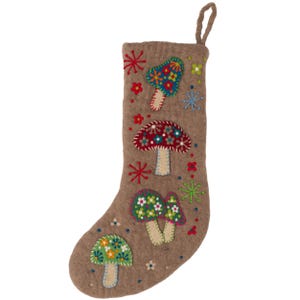 May include: A tan felt Christmas stocking featuring embroidered mushroom designs in blue, red, and green with floral patterns. The stocking includes a hanging loop at the top and is decorated with small stitched starbursts and flower accents throughout.
