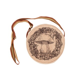 May include: A round, beige canvas bag with a brown strap. The bag features a brown line drawing of a mushroom with a snail on top, surrounded by flowers and leaves.