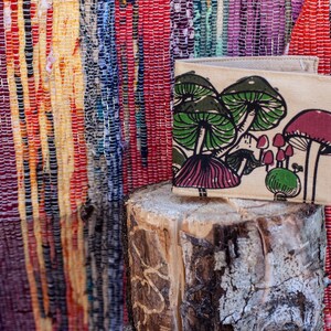 Pu&ograve; includere: Un portafoglio in tela beige con una stampa di funghi. I funghi sono verdi, rosa e bianchi. Il portafoglio &egrave; appoggiato su un tronco di legno.