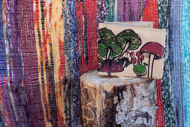 Pu&ograve; includere: Un portafoglio in tela beige con una stampa di funghi. I funghi sono verdi, rosa e bianchi. Il portafoglio &egrave; appoggiato su un tronco di legno.