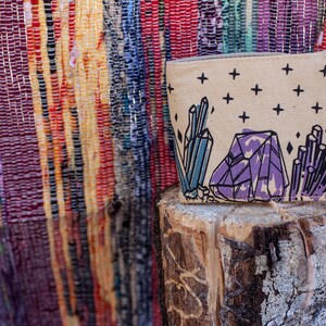 Pu&ograve; includere: Una pochette in tela beige con un designo di cristallo viola, blu e nero. La pochette &egrave; appoggiata su un tronco di legno.