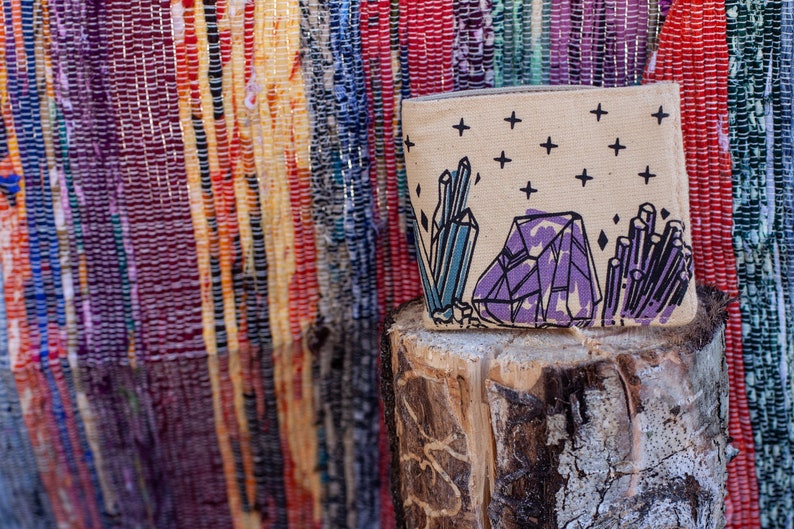 Pu&ograve; includere: Una pochette in tela beige con un designo di cristallo viola, blu e nero. La pochette &egrave; appoggiata su un tronco di legno.