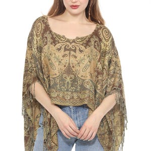 Può includere: Un poncho marrone e beige con motivo paisley e finiture con frange. Il poncho ha uno scollo tondo e maniche corte. Il tessuto presenta un design dettagliato con motivi floreali e a volute. La modella indossa jeans blu.