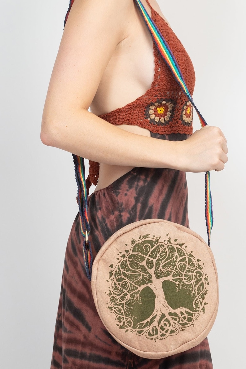 May include: A round, beige crossbody bag with a green tree of life design. The bag has a rainbow striped strap.