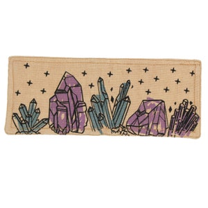 Pu&ograve; includere: Un cerotto in tessuto beige con un disegno di cristalli viola e blu su uno sfondo beige con stelle nere.