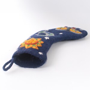 Op de afbeelding: Een donkerblauwe kerstkous van gevilt wol met geborduurde hemelse motieven, waaronder een gele zon, een halve maan en een kleurrijke vlinder. De kous heeft een klein lusje aan de bovenkant om op te hangen en kleine witte steraccenten.