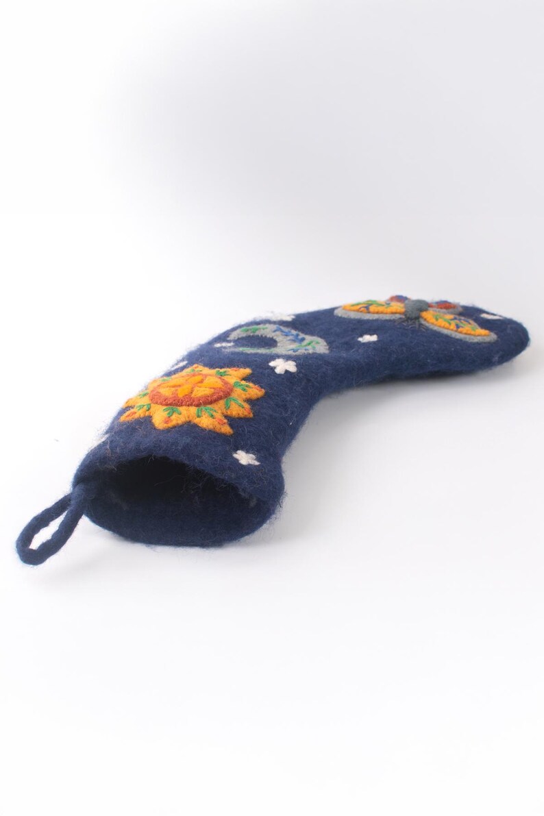 Op de afbeelding: Een donkerblauwe kerstkous van gevilt wol met geborduurde hemelse motieven, waaronder een gele zon, een halve maan en een kleurrijke vlinder. De kous heeft een klein lusje aan de bovenkant om op te hangen en kleine witte steraccenten.