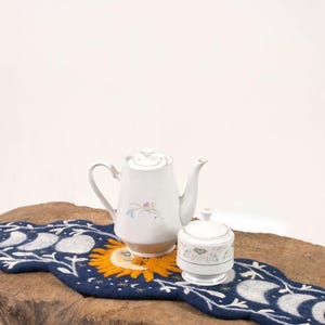 May include: A white porcelain teapot and sugar bowl sit on a navy blue felt table runner with a sun and moon design. The teapot has a gold base and floral accents. The sugar bowl has a lid and floral pattern.