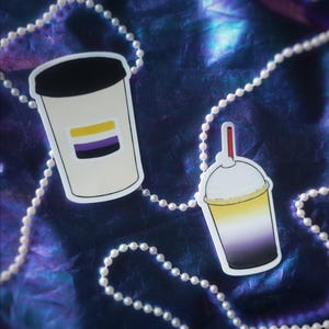 Puede incluir: Dos pegatinas con un diseño de la bandera del orgullo no binario. Una pegatina es una taza de café blanca con tapa negra y una franja amarilla y morada. La otra pegatina es una taza degradada con pajita roja.