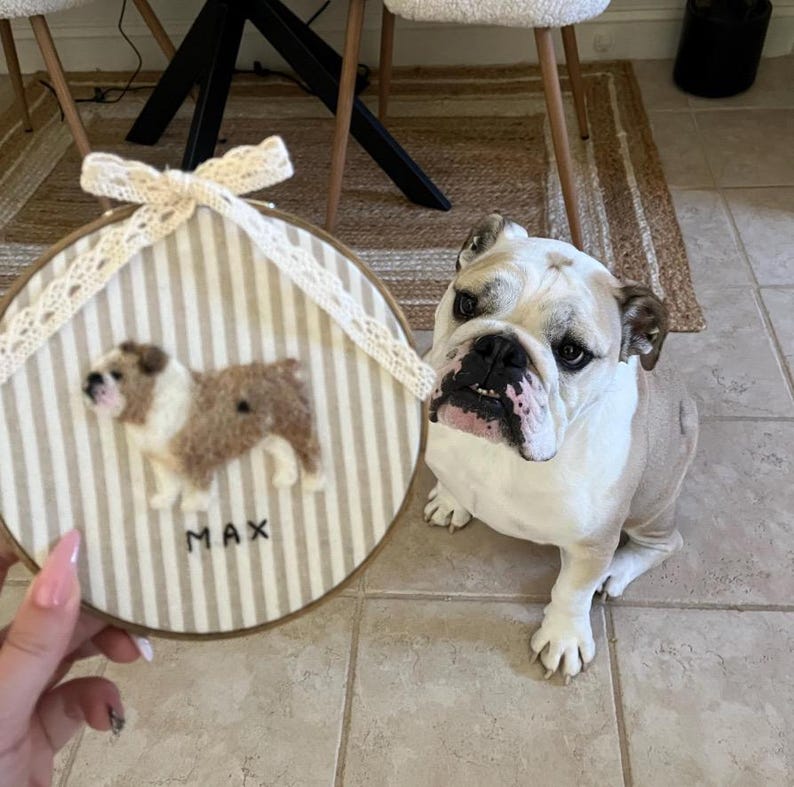 May include: A round, framed artwork featuring a felted bulldog with brown and white fur, set against a striped background. The name "MAX" is embroidered below the dog. A real bulldog sits next to the artwork.