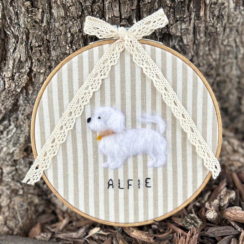 May include: A round, wooden embroidery hoop featuring a white dog with the name "ALFIE" stitched below. The background is striped beige fabric. A cream-colored lace bow and trim adorn the top.