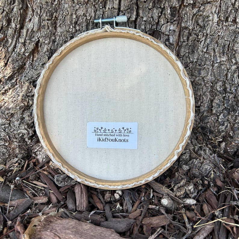 May include: A round embroidery hoop with a cream-colored fabric backing. The wooden hoop is secured with a metal clasp. A white label with the text "Hand stitched with love iKidYouKnots" is affixed to the fabric.