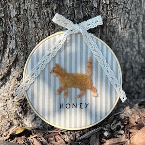 May include: A round, beige and white striped embroidery hoop featuring a felted orange cat. The word "HONEY" is embroidered below the cat. A cream-colored lace bow is attached at the top.