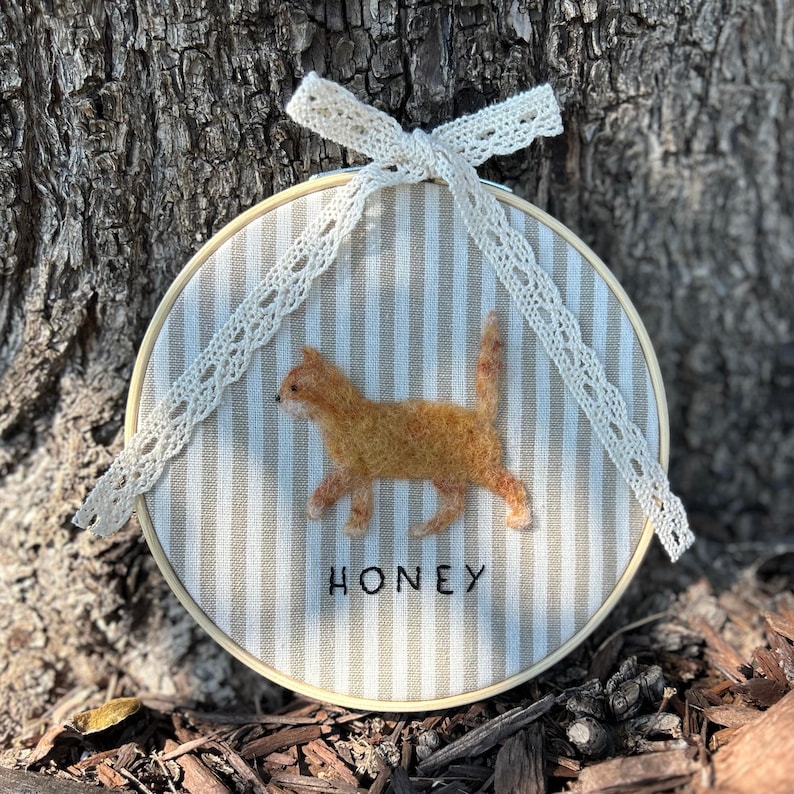 May include: A round, beige and white striped embroidery hoop featuring a felted orange cat. The word "HONEY" is embroidered below the cat. A cream-colored lace bow is attached at the top.