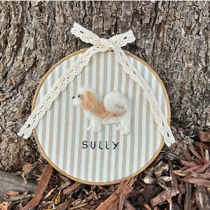 May include: A round, framed embroidery featuring a fluffy, tan and white dog with the name "SULLY" stitched below. The background is striped beige and white fabric, with a cream lace bow at the top.