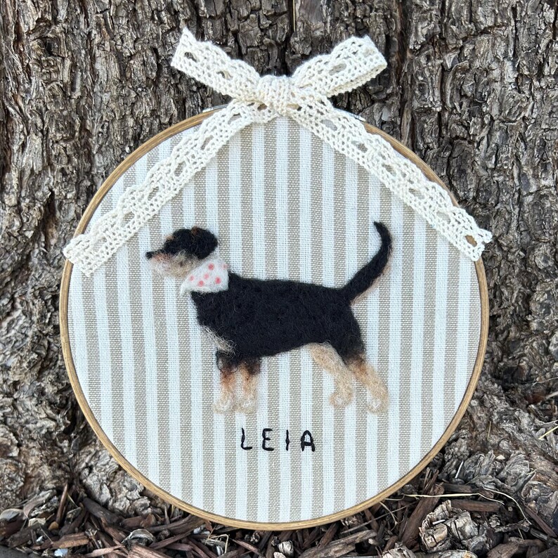 May include: A handmade dog portrait in an embroidery hoop. The dog is black, tan, and white, wearing a pink and white polka dot bandana. The background is striped, and the dog's name, "LEIA," is written below. A cream-colored lace bow adorns the top.