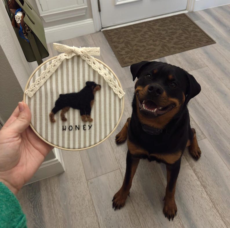 May include: A round, framed embroidery hoop featuring a Rottweiler dog silhouette on a striped background. The dog's name, "HONEY," is stitched below. The hoop is adorned with a lace bow. A Rottweiler dog is sitting next to the hoop.