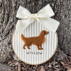 May include: A round, wooden embroidery hoop featuring a brown dog silhouette on a striped background. The dog's name, "WILLOW," is stitched below. A cream-colored bow is attached at the top, creating a decorative piece.