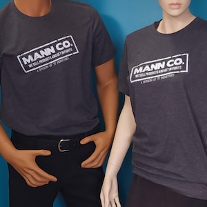 May include: A charcoal gray short-sleeve t-shirt featuring a white rectangular graphic with the text MANN CO. WE SELL PRODUCTS AND GET IN FIGHTS A DIVISION OF TF INDUSTRIES. The shirt has a crew neckline and a standard fit on the torso.