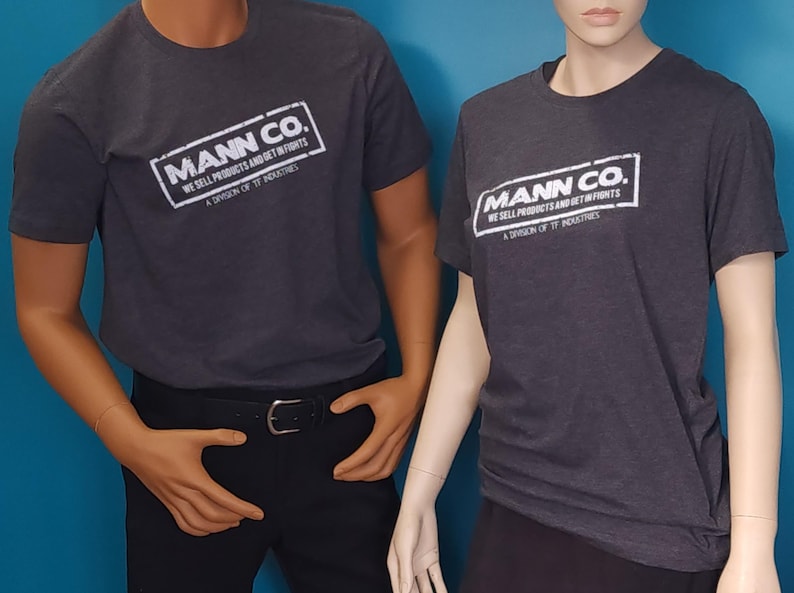 May include: A charcoal gray short-sleeve t-shirt featuring a white rectangular graphic with the text MANN CO. WE SELL PRODUCTS AND GET IN FIGHTS A DIVISION OF TF INDUSTRIES. The shirt has a crew neckline and a standard fit on the torso.