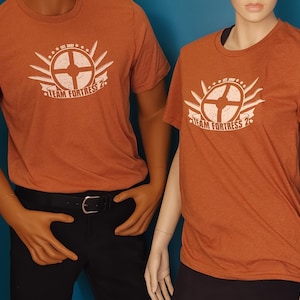 May include: Two short-sleeved crew neck t-shirts in a heathered burnt orange color. Each shirt features a white graphic print on the chest displaying a circular emblem with radiating lines, stars, and the text "TEAM FORTRESS 2" with "EST. 2007".