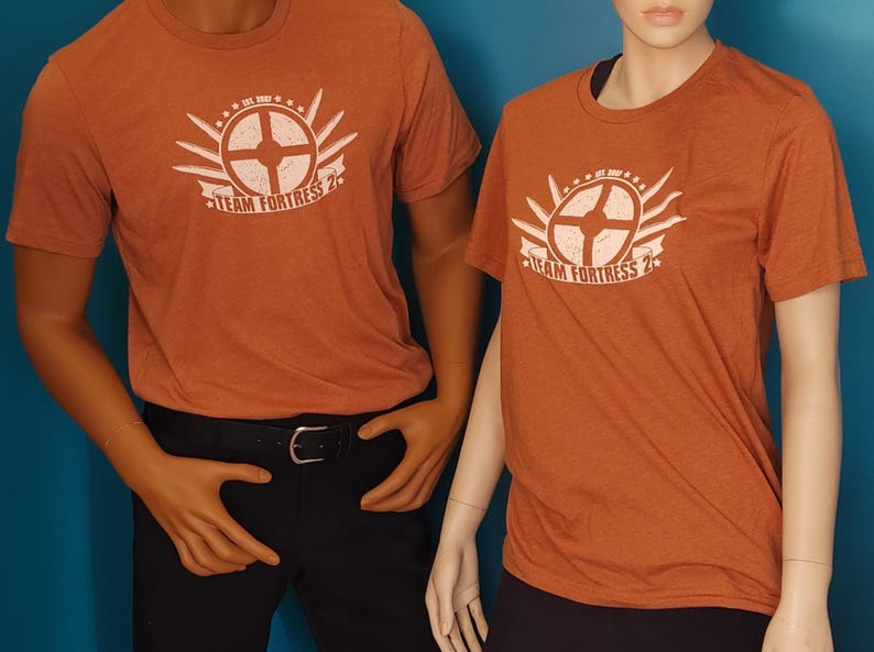 May include: Two short-sleeved crew neck t-shirts in a heathered burnt orange color. Each shirt features a white graphic print on the chest displaying a circular emblem with radiating lines, stars, and the text "TEAM FORTRESS 2" with "EST. 2007".