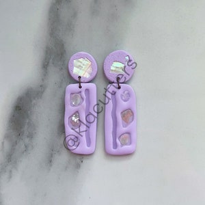 May include: A pair of lavender polymer clay earrings with iridescent accents. Each earring features a rectangular shape with three small, iridescent circles embedded in the clay. The top of each earring is a round, lavender circle with a larger iridescent circle embedded in the clay.