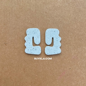 May include: A pair of light blue abstract shaped earrings made of speckled terrazzo. The earrings are in the shape of a rounded rectangle with a cut-out in the middle. The earrings are shown on a brown background. BUYKLA.COM is printed in white text below the earrings.