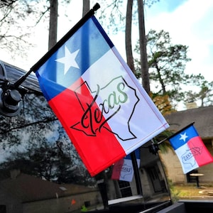 May include: Texas state flag with a white star on a blue background, a white stripe, and a red stripe. The flag has the word "Texas" in a cursive font and a black outline of the state.