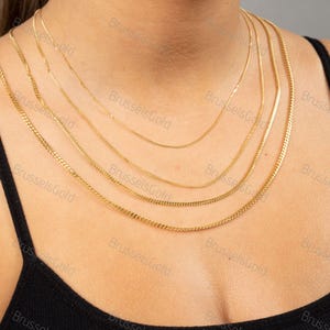May include: Five gold chains of different lengths and widths layered on a person's neck. The chains are all made of a shiny gold metal.
