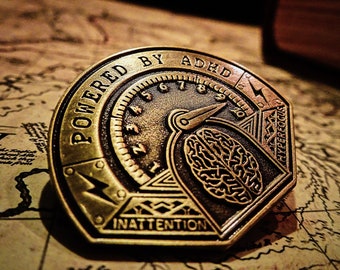 May include: A gold-toned metal pin with a stylized brain design and the text "POWERED BY ADHD" and "INATTENTION" on the bottom. The pin also features a stylized speedometer with the text "HYPERFOCOUS" on the right side.