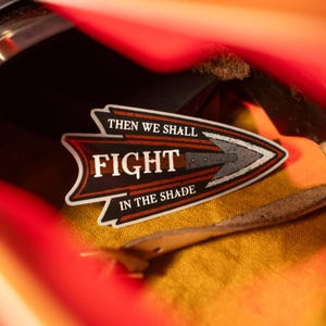 Puede incluir: Una pegatina de flecha blanca con el texto "Then we shall fight in the shade" y un gráfico de cuchillo estilizado. La pegatina está sobre una superficie de cuero marrón.