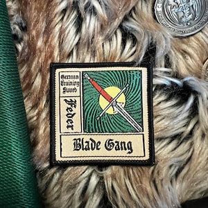 Puede incluir: Un parche bordado de color beige y negro con un fondo verde y blanco. El parche presenta una espada con mango rojo y el texto "German Training Sword Feder Blade Gang".