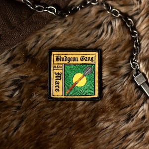 Könnte beinhalten: Ein braunes und schwarzes gesticktes Patch mit dem Text "Bludgeon Gang" und "2.5 lb Mace" in einem gelben Kasten. Das Patch hat einen grünen Hintergrund mit einer gelben Sonne und einer braunen und schwarzen Keule.