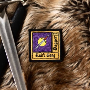 May include: A black and yellow embroidered patch with a purple background. The patch features a stylized dagger with a yellow sun and the text "Golden Dagger Knife Gang".