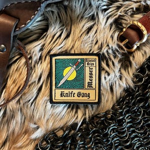 May include: A gold and black embroidered patch with a green background. The patch features a stylized sword with a yellow sun behind it. The text on the patch reads "Riveted Grips Messer Knife Gang".