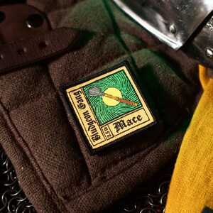 May include: A brown and green embroidered patch with a yellow background. The patch features a stylized illustration of a mace with the text "Bludgeon Gang" and "Mace" on it.