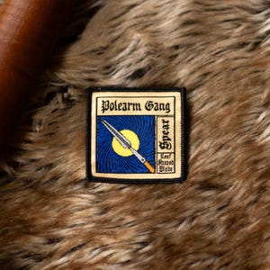 Puede incluir: Un parche bordado negro y amarillo con el texto "Polearm Gang" y "Spear" con un dibujo de una lanza con una hoja en forma de hoja.