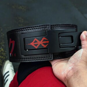 Op de afbeelding: Zwarte gewichthefband met een rode afbeelding en Japanse tekst. De riem heeft een zwarte gesp. De persoon die de riem vasthoudt, draagt een rode korte broek en witte sneakers.