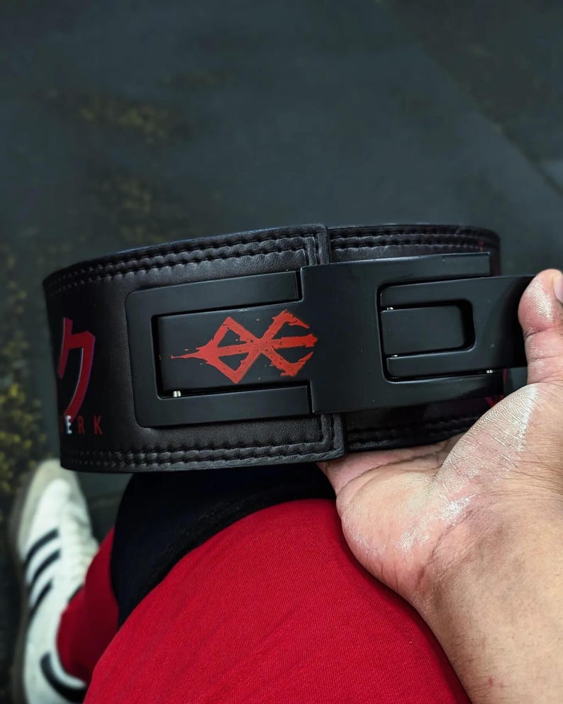 Op de afbeelding: Zwarte gewichthefband met een rode afbeelding en Japanse tekst. De riem heeft een zwarte gesp. De persoon die de riem vasthoudt, draagt een rode korte broek en witte sneakers.