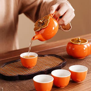 May include: An orange ceramic teapot with a flower-shaped lid pours tea into a matching cup. The set includes a small lidded container and several cups, all in the same vibrant orange colour. The tea set is on a woven tray.