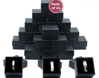 May include: A pack of 24 black ring boxes, each with a black velvet interior. The boxes are stacked in a pyramid shape. Three boxes are shown with rings inside, one with a gold band, one with a silver band, and one with a silver band with clear stones.