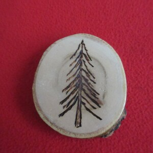 Pode incluir: Um ornamento redondo de madeira com um desenho de pinheiro queimado sobre um fundo vermelho.