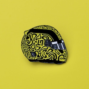 Könnte beinhalten: Gelb-schwarzer Emaille-Pin in Helmform. Der Pin hat ein abstraktes schwarz-gelbes Design mit schwarzem Visier und Details. Der Pin ist vor einem einfarbigen gelben Hintergrund platziert.