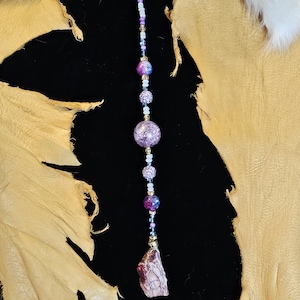 May include: A long beaded necklace with a purple and white color scheme. The necklace features a large purple and white crystal bead and a smaller purple crystal bead. The necklace ends with a large, irregular shaped purple and white crystal.