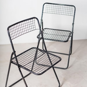 May include: Two black metal folding chairs with a wire mesh seat and back. The chairs are in a white room.